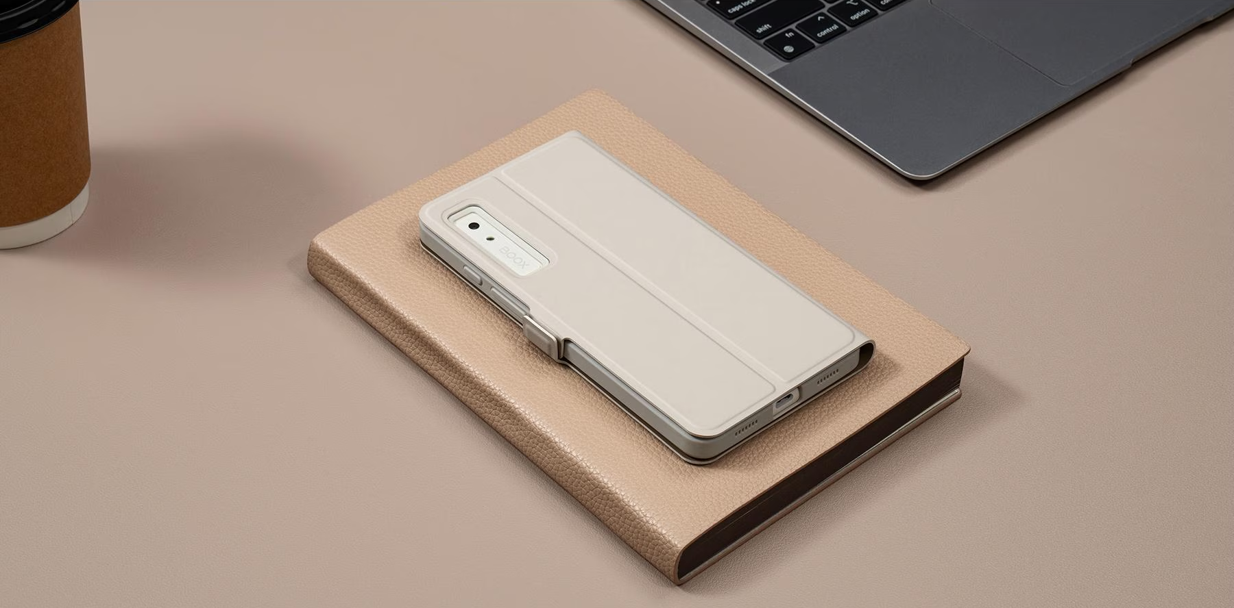 Smartphone in cremefarbenem Case auf geschlossenem Buch, Laptop und Kaffeetasse in der Nähe.