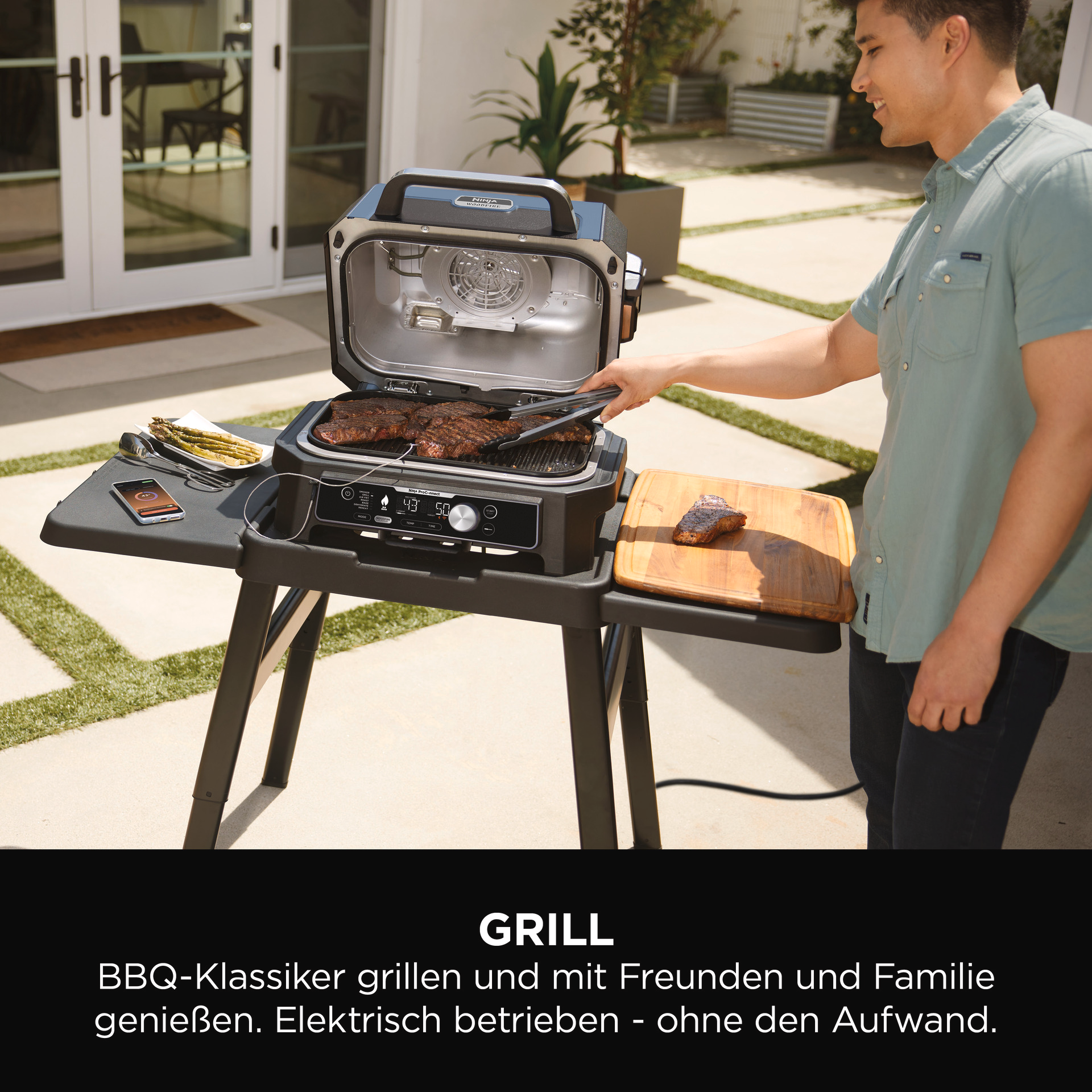 Mann grillt Steaks auf einem schwarzen Ninja-Grill. Ein Holzschneidebrett und Telefon sind in der Nähe. Text: GRILL.