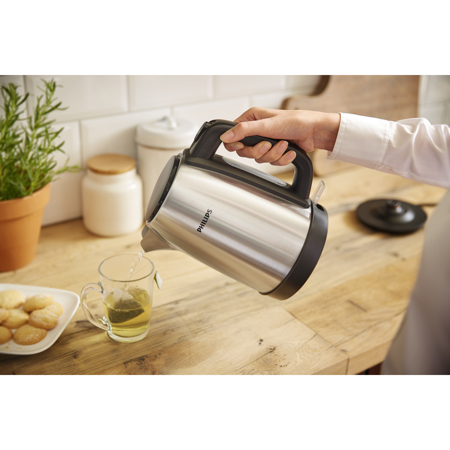 Een hand giet water uit een zilveren Philips waterkoker in een glas thee op hout.