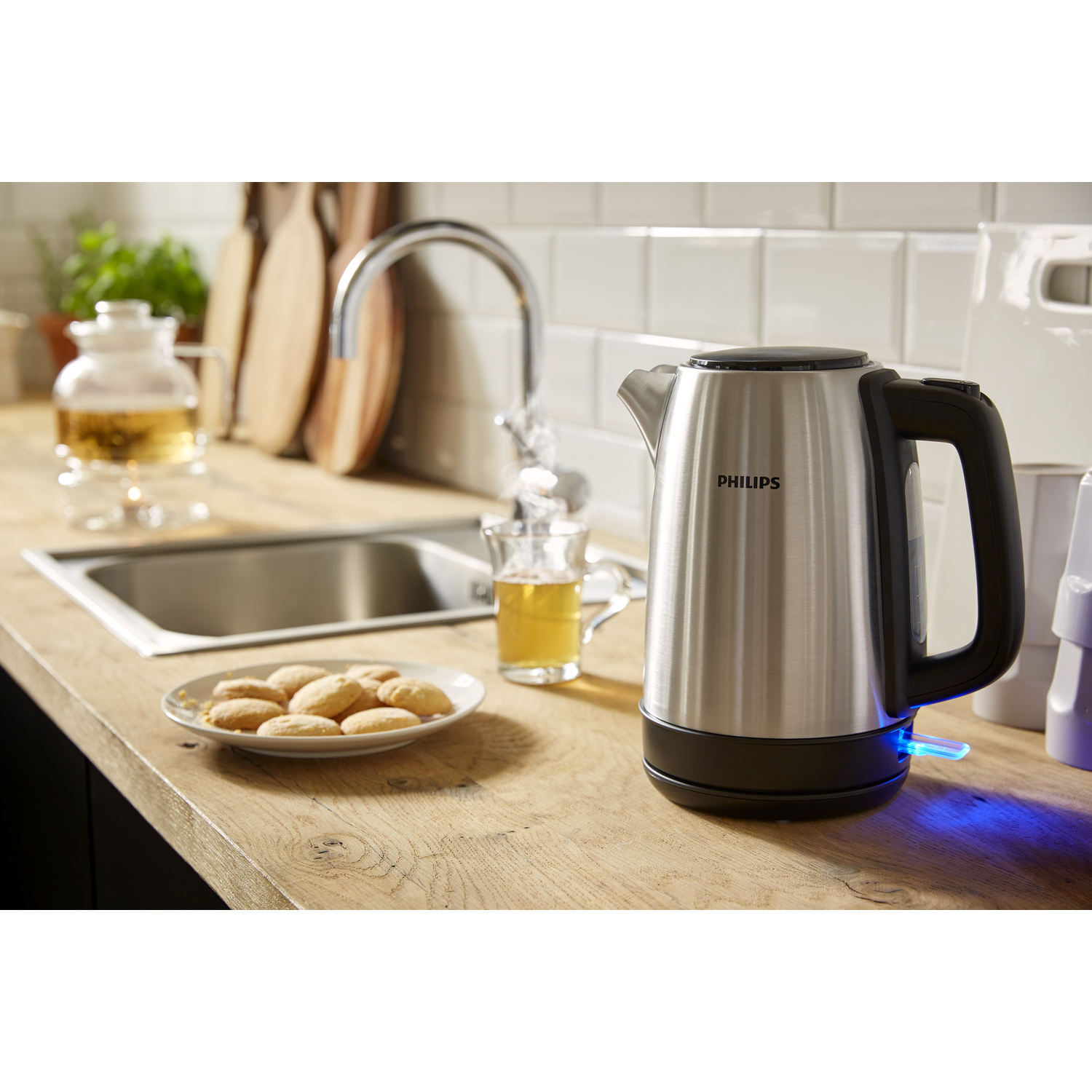 Zilveren Philips waterkoker op houten oppervlak met kopje thee, koekjes en een gootsteen.