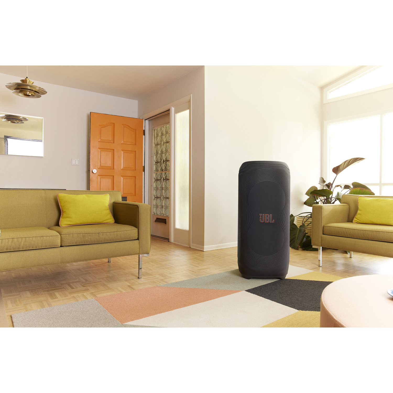 Een woonkamer met een gele bank, een zwarte luidspreker en een kleurrijk tapijt. De luidspreker heeft een JBL logo.