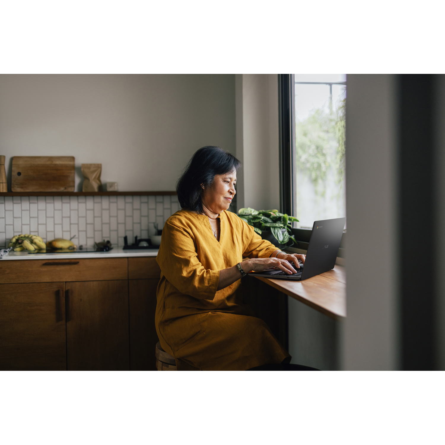 Vrouw in gele jurk gebruikt laptop aan houten tafel bij raam. Houten kasten op de achtergrond.
