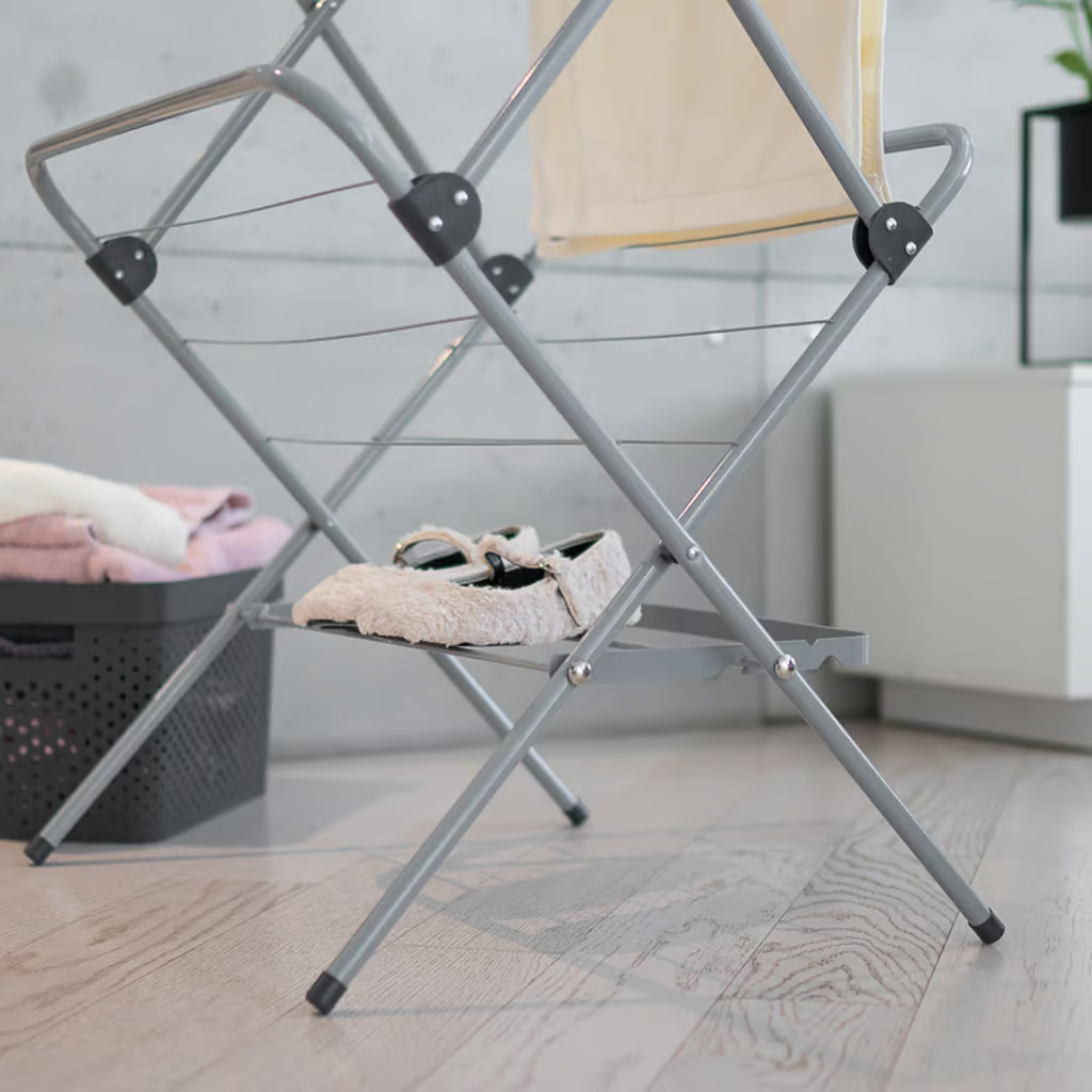 Szary stojak do suszenia ubrań z ubraniami i butami. W tle czarny kosz i szafka.