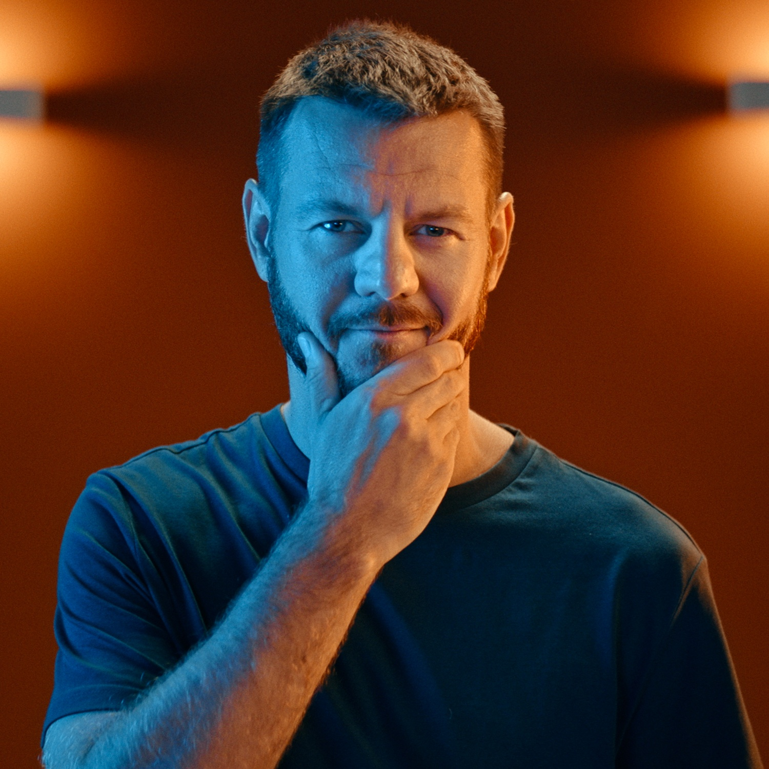 Un uomo con la barba si tocca il mento, guardando la fotocamera sotto luci arancioni e blu.