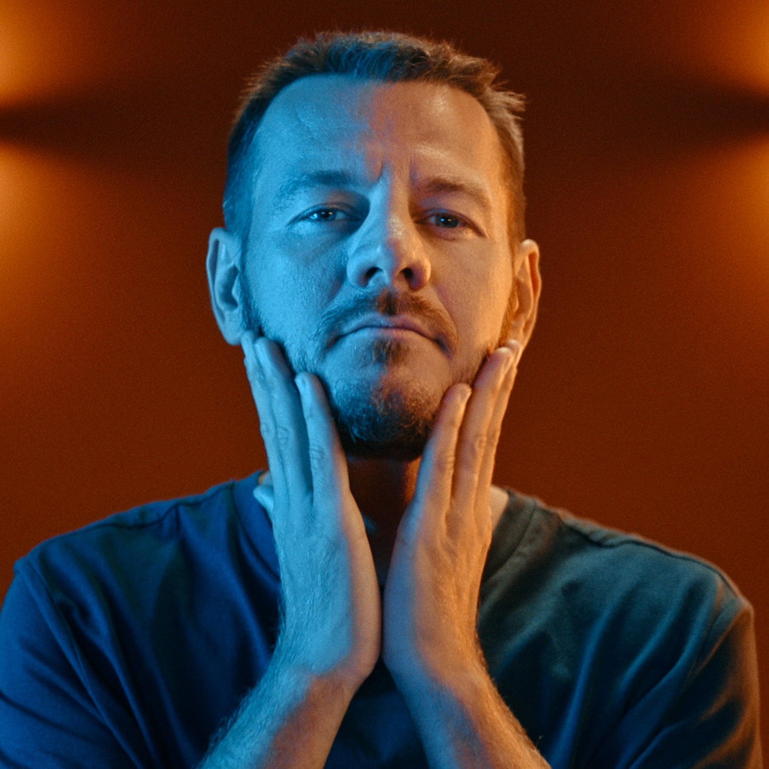 Un uomo si tiene il viso con entrambe le mani. Lo sfondo è arancione e la luce blu sul suo viso.