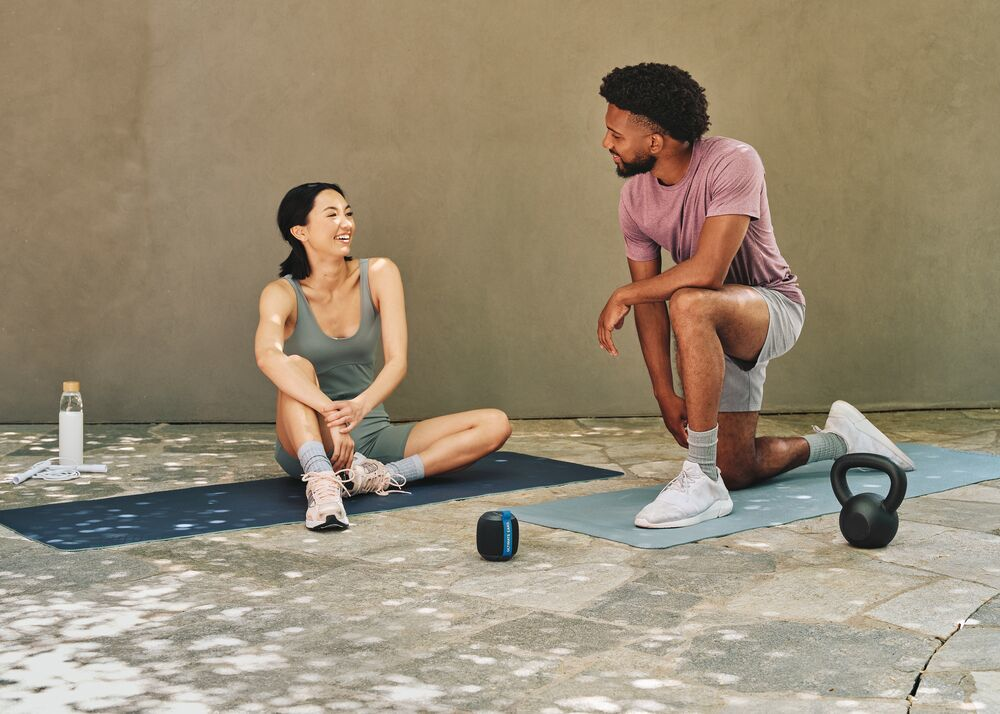 Ein Mann und eine Frau auf Yogamatten, lächelnd, mit Lautsprecher und Kettlebell in der Nähe.