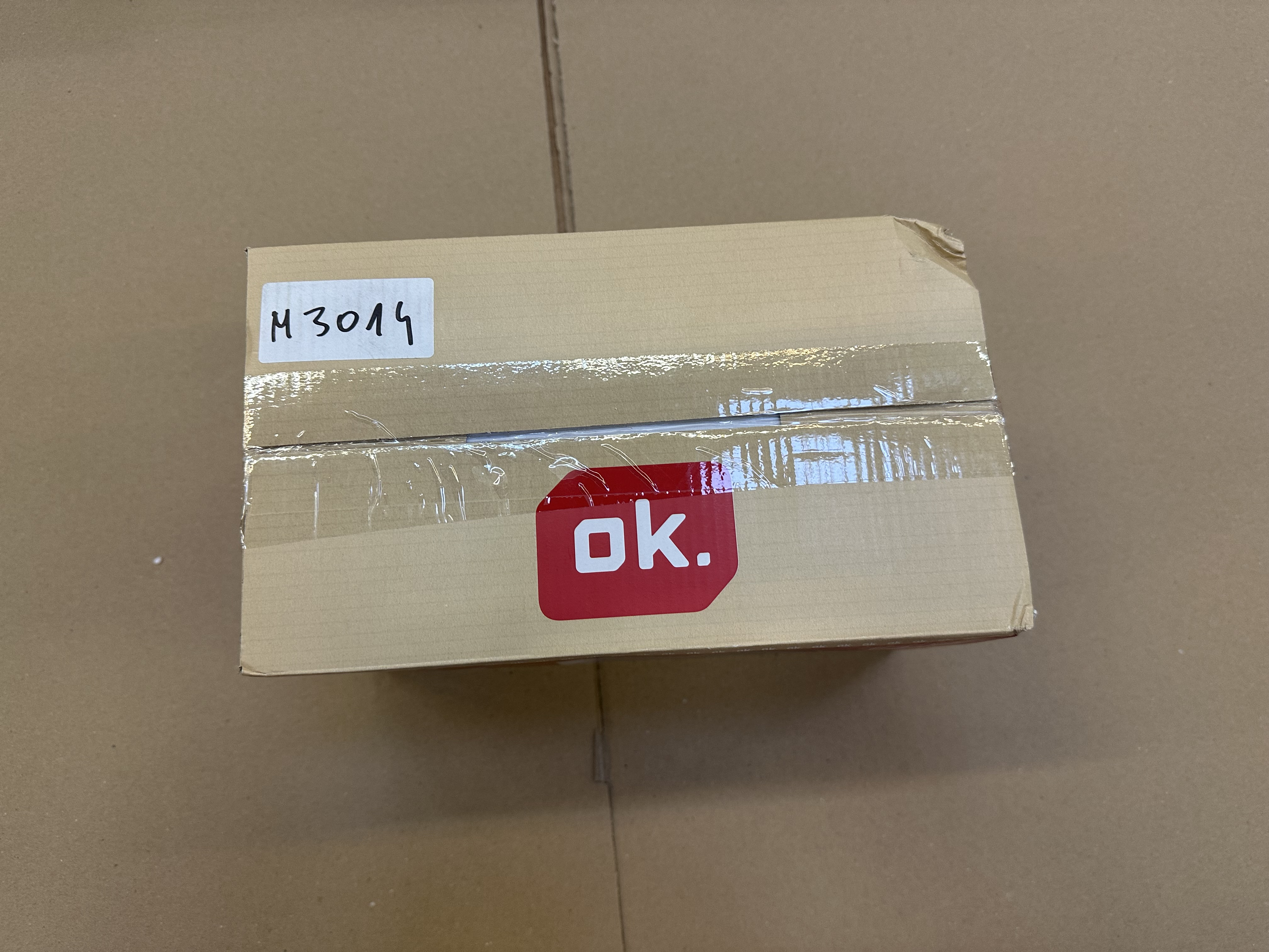 Kartonowe pudełko z taśmą i naklejką „ok.”. Na pudełku znajduje się odręczna etykieta „M3014”.