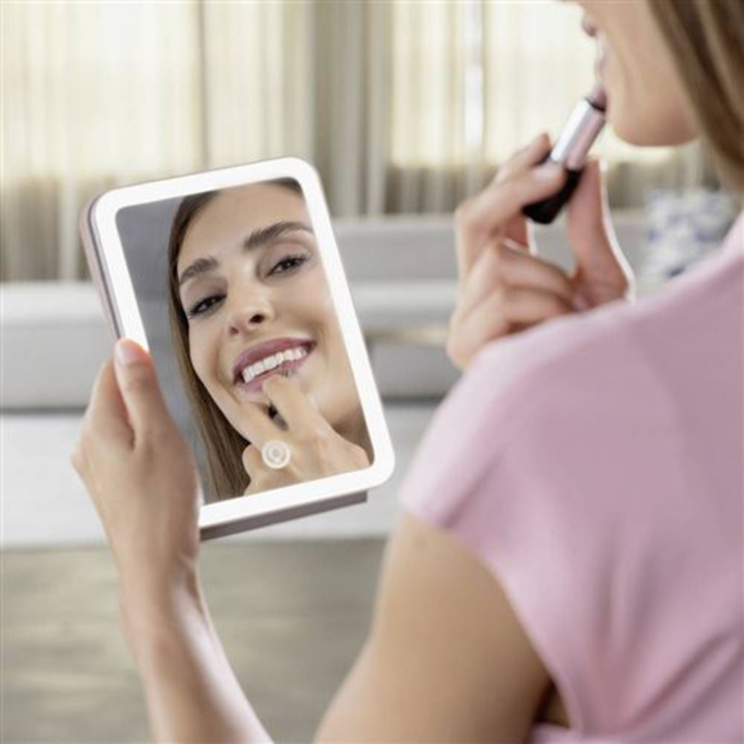 A woman applies lipstick in a lighted mirror. Reflection shows her smiling and touching her lip.