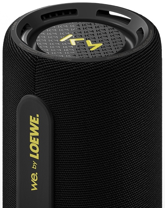 Czarny, cylindryczny głośnik z żółtym tekstem i czarną górą. Tekst brzmi "we. by LOEWE".