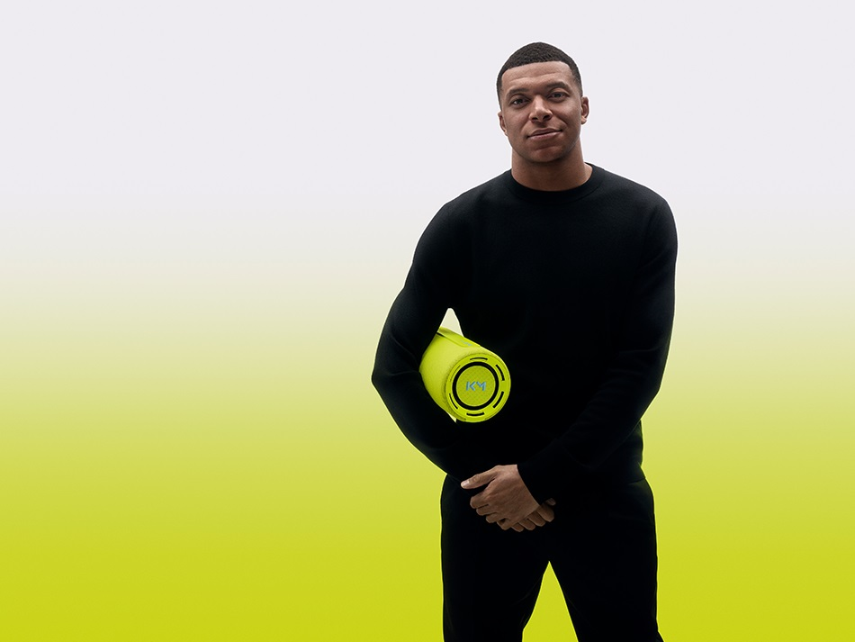 Kylian Mbappe trzyma zielony wałek. Żółte i białe tło. Uśmiechnięty.