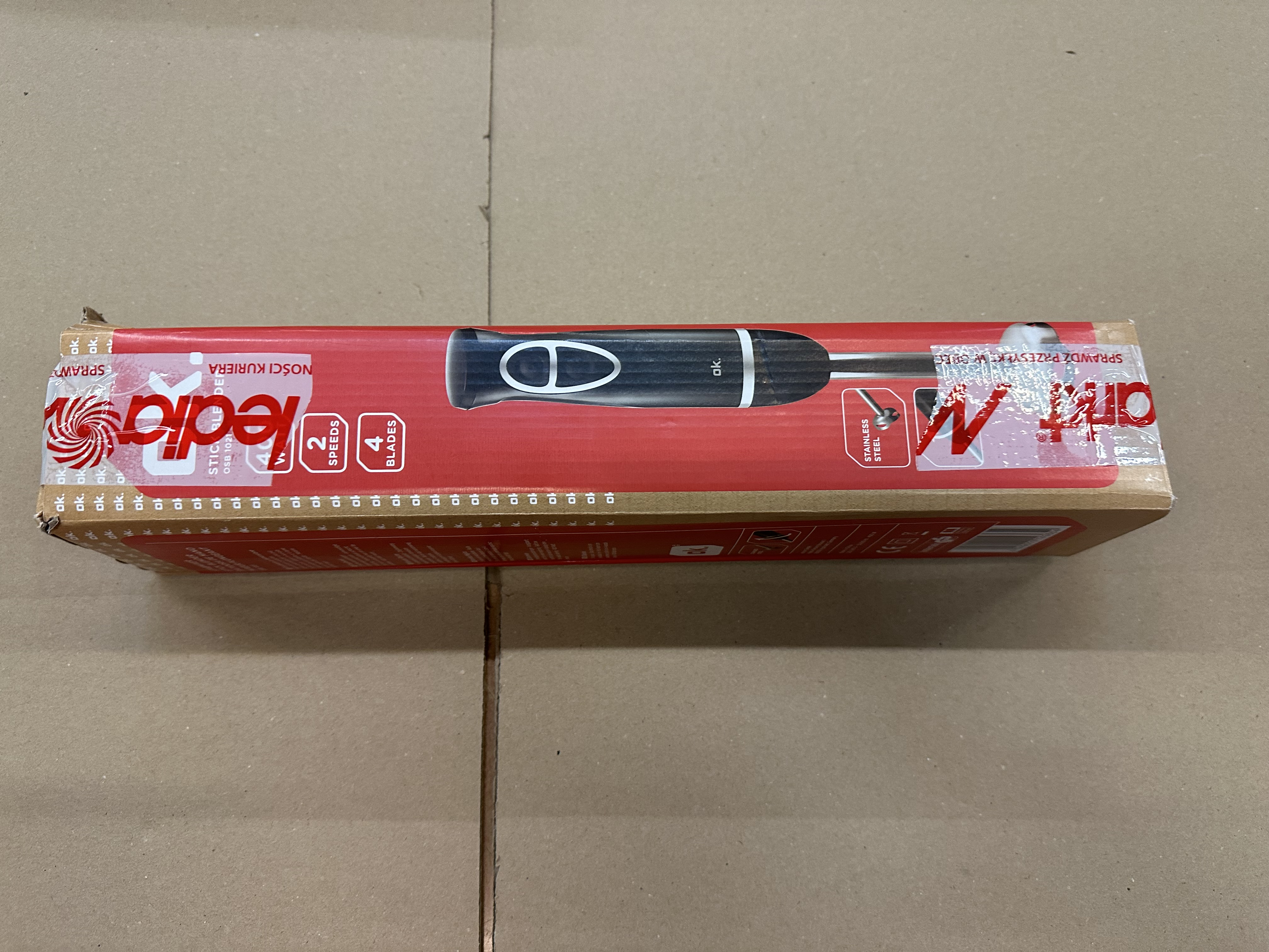 Pudełko blendera na brązowej powierzchni. Pudełko jest czerwone z czarnymi detalami i tekstem.