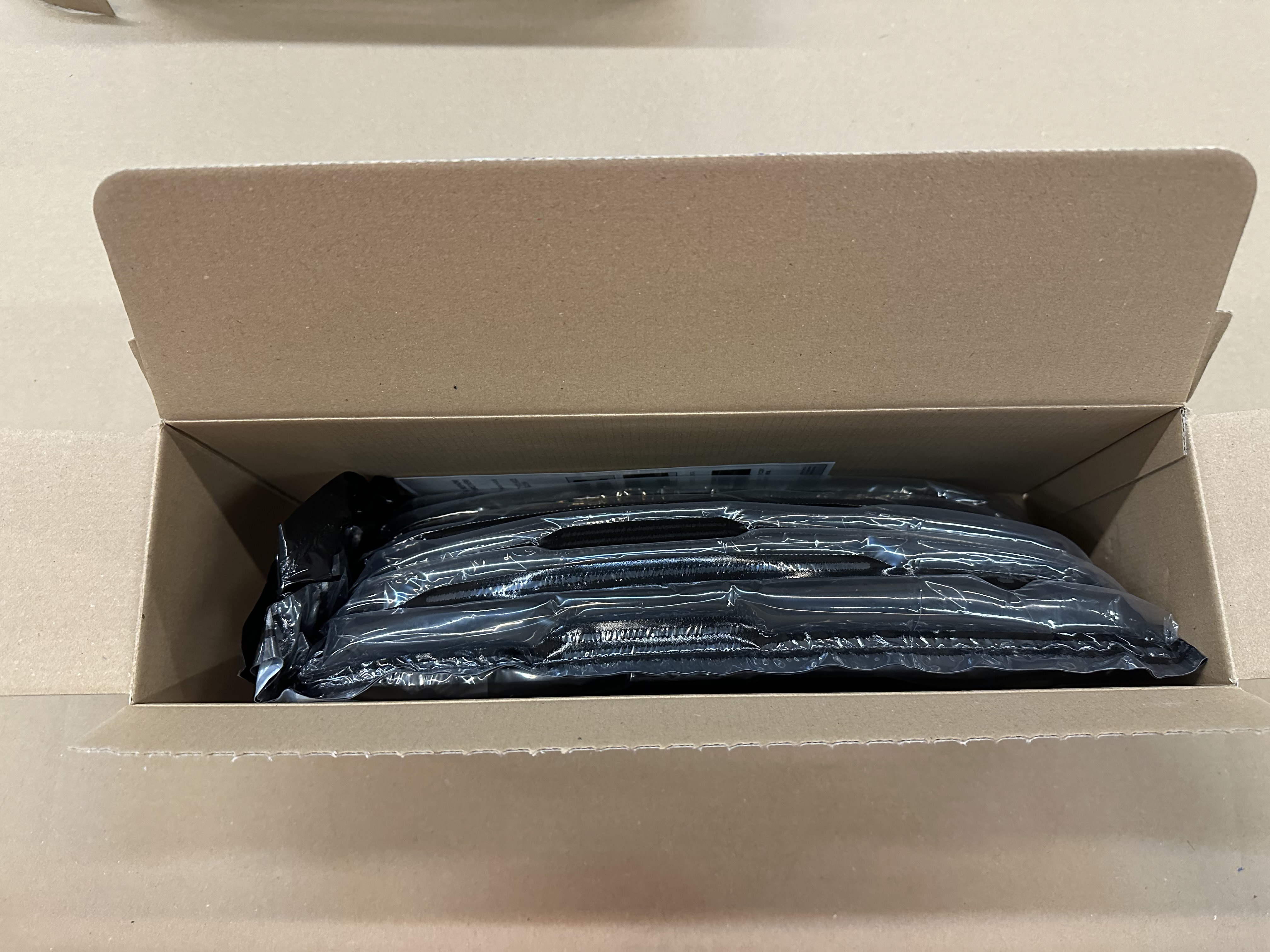 Otwarty karton. Wewnątrz widoczny czarny przedmiot owinięty w plastik.