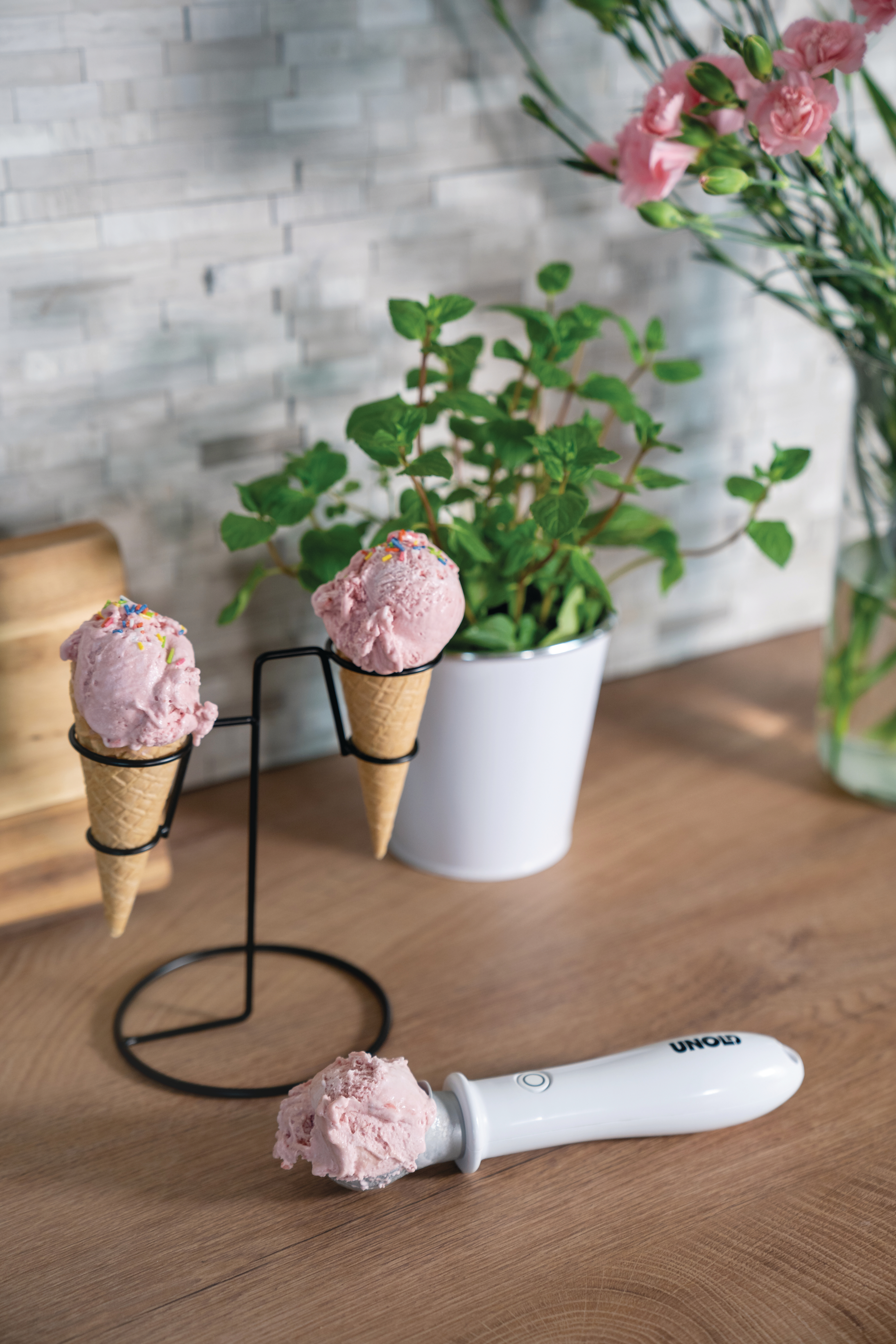 Eistüten auf einem Ständer, Eisportionierer und ein Topf mit Minze auf einem Holztisch.