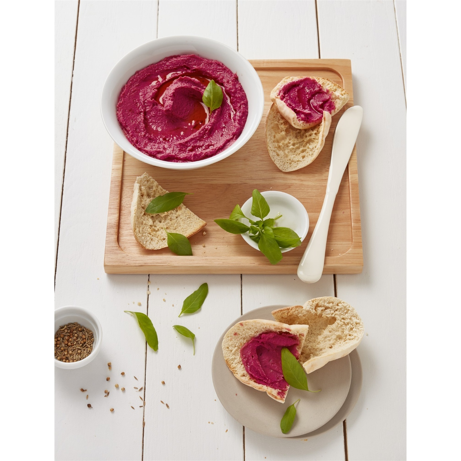Hummus di barbabietole con pane, basilico e olio d'oliva, su tagliere di legno e superficie bianca.