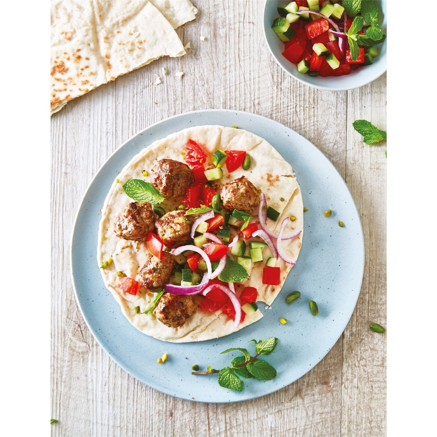 Un piatto con un wrap contenente polpette, pomodori, cetrioli, cipolla rossa e menta. Pane piatto e una ciotola.