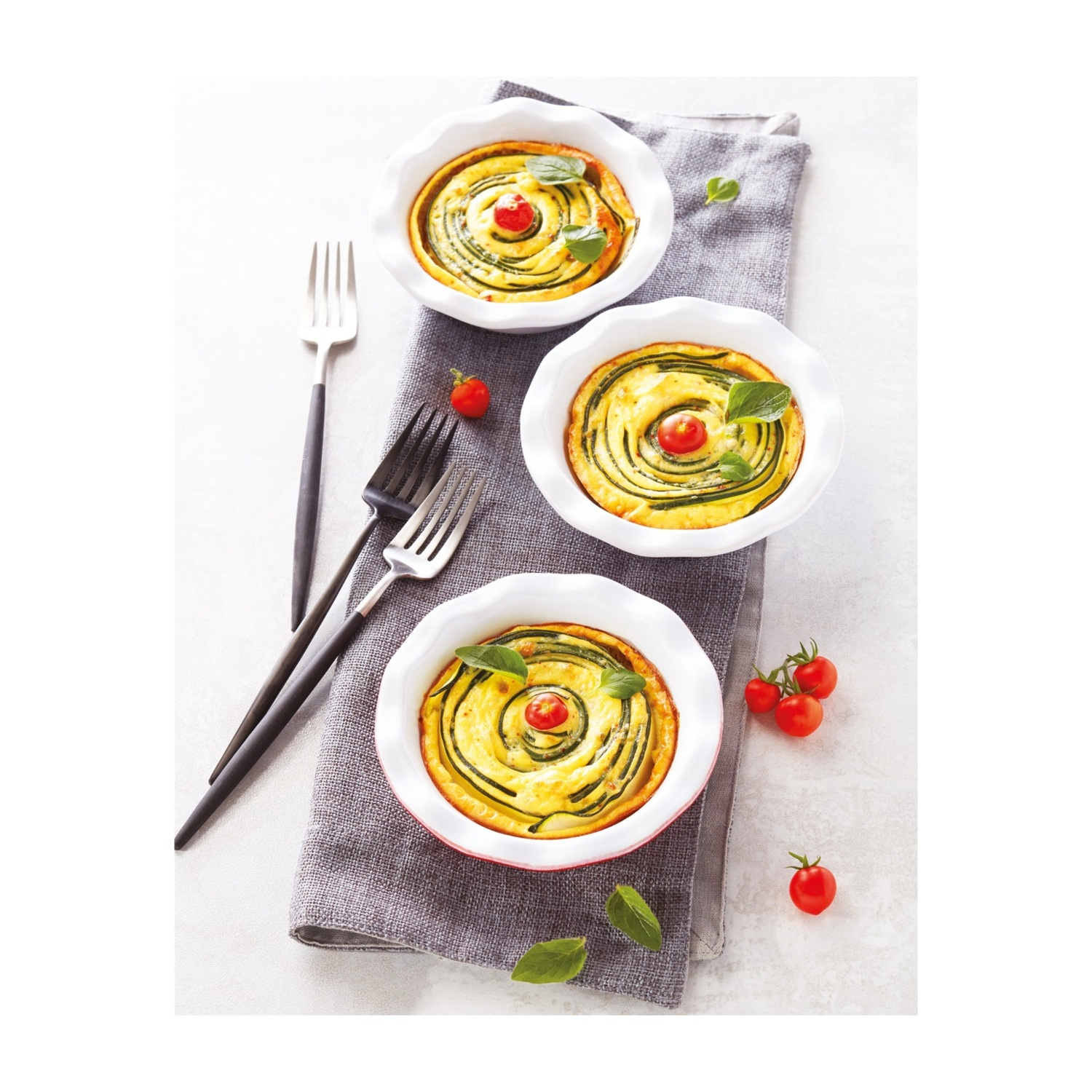Tre involtini al forno di zucchine e formaggio con pomodorini e basilico su un panno grigio.