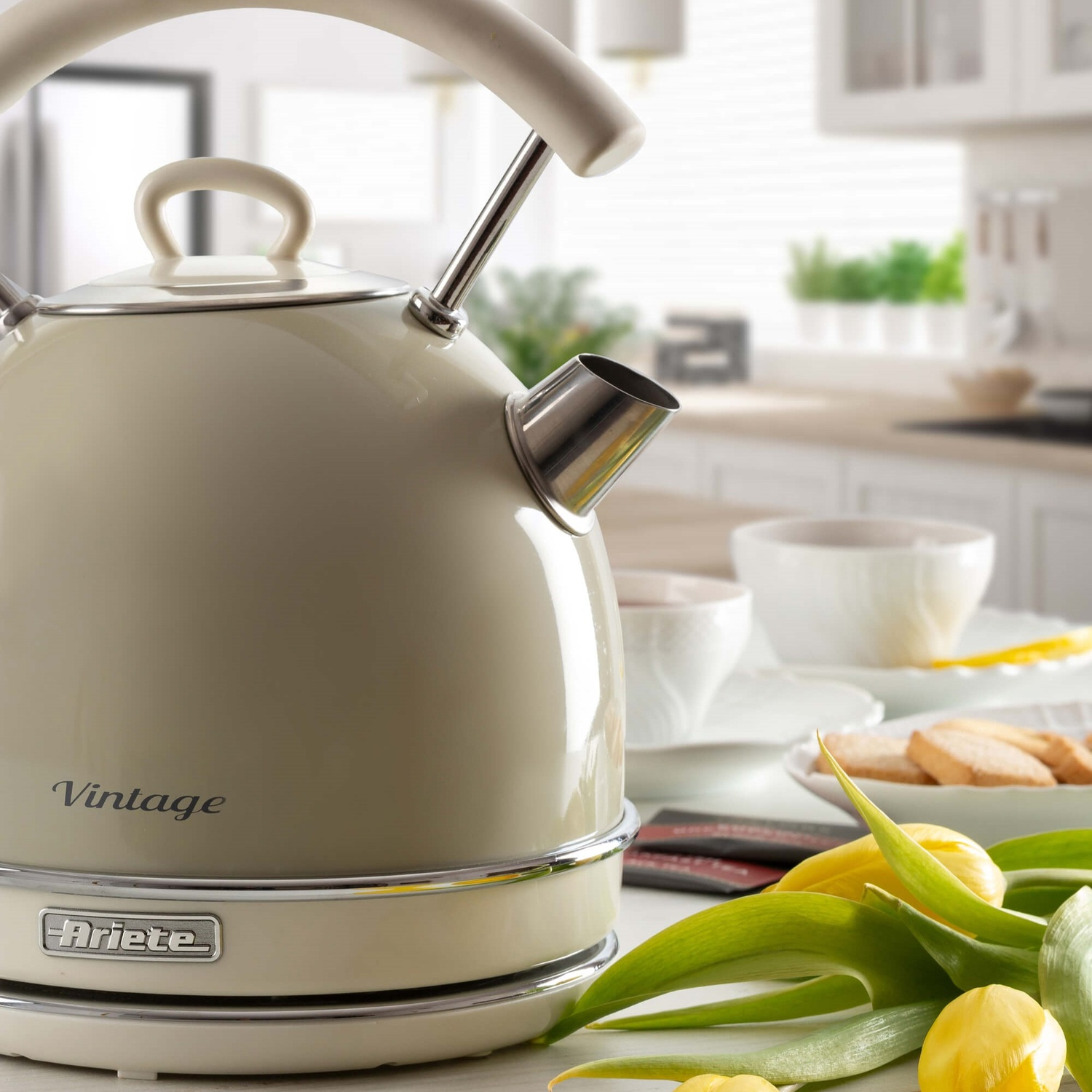Bollitore color crema con manico e beccuccio. È su una base, vicino a una tazza e fiori.