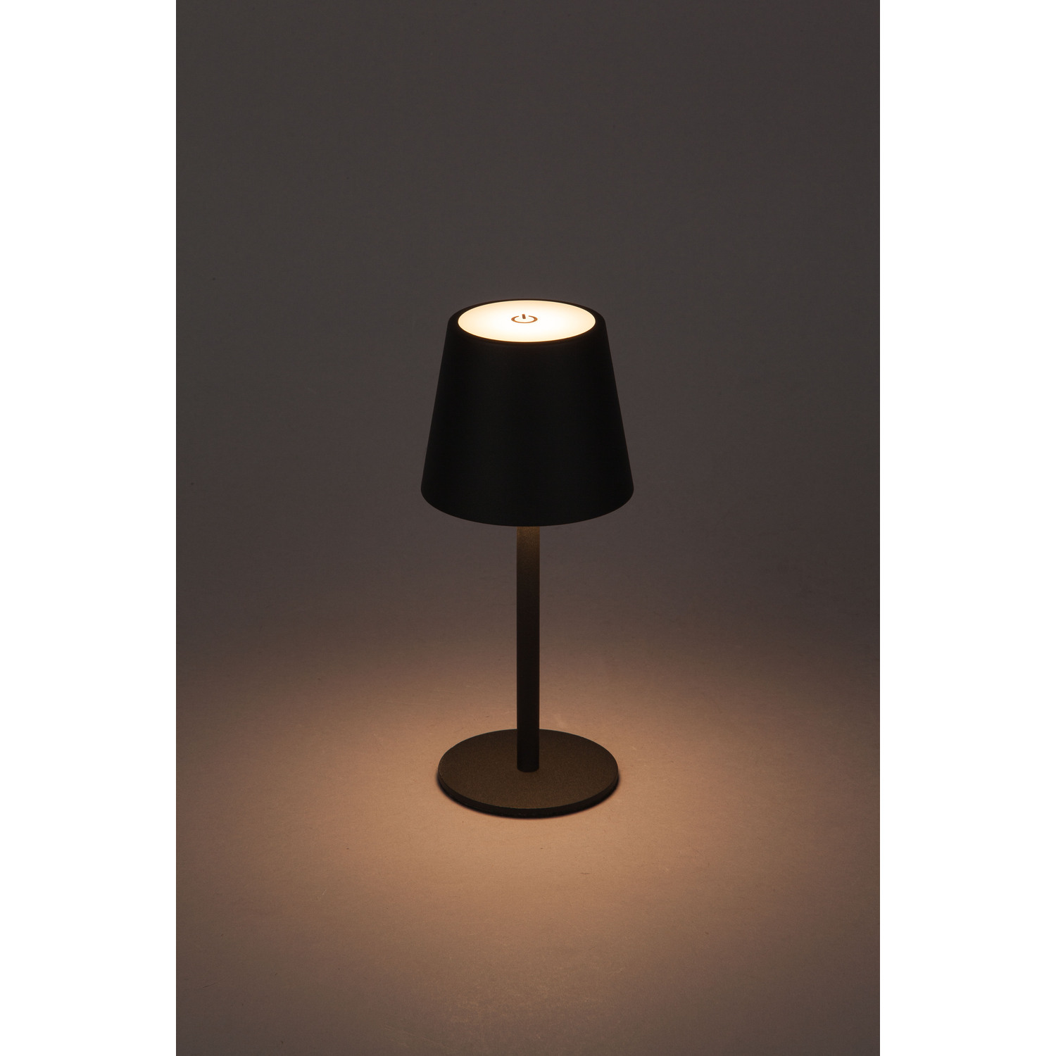 A black table lamp with a cylindrical shade, lit against a dark backdrop.