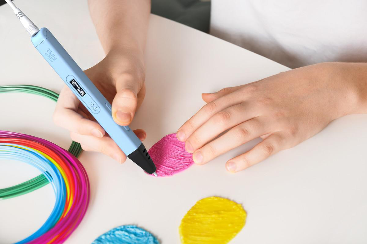 Eine Person verwendet einen blauen 3D-Stift, um eine rosa Form auf einer weißen Oberfläche zu erstellen. Andere Kunststofffarben sind sichtbar.
