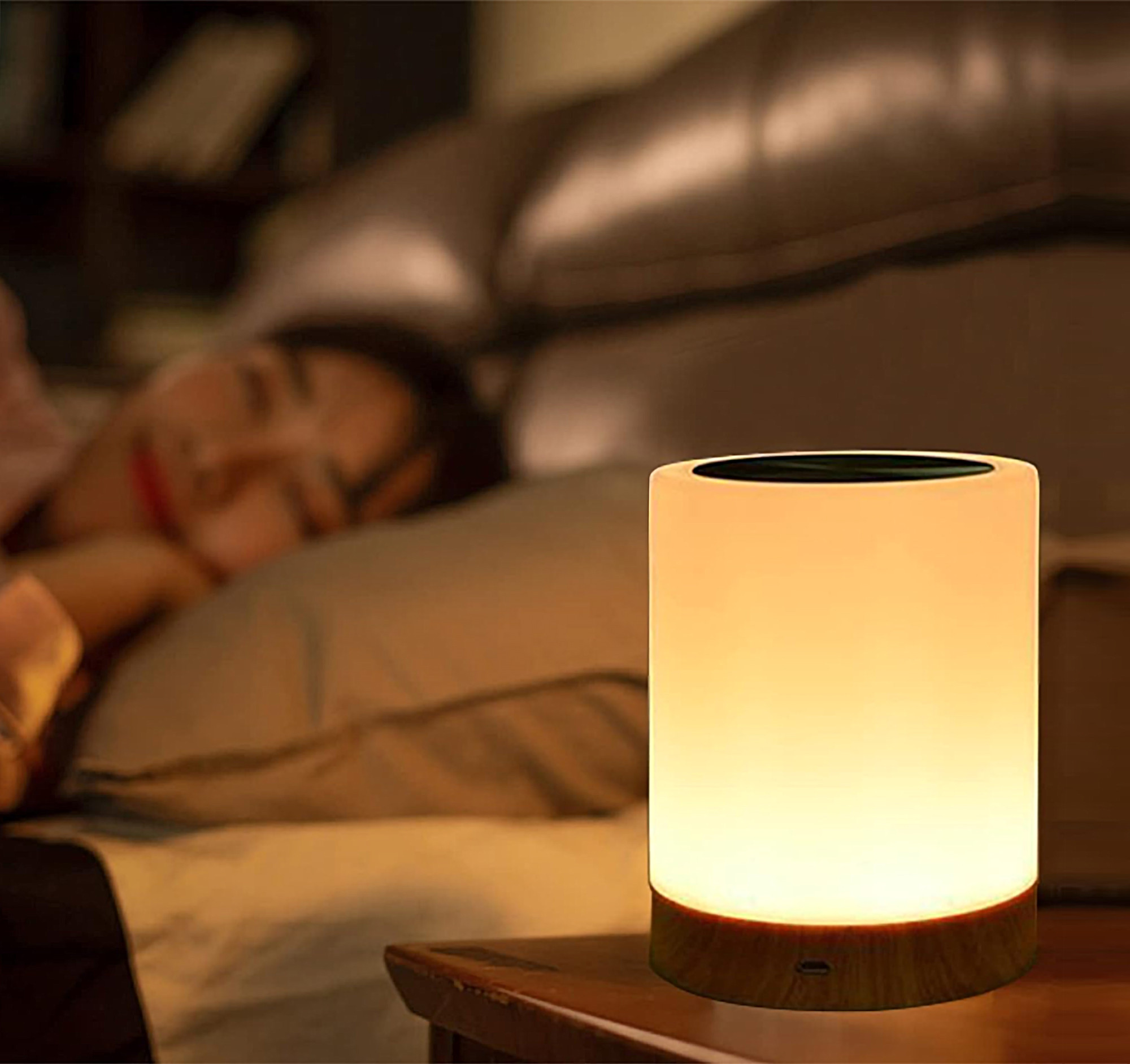 Eine Nachttischlampe mit Holzsockel leuchtet auf einem Nachttisch. Im Hintergrund schläft eine Frau.