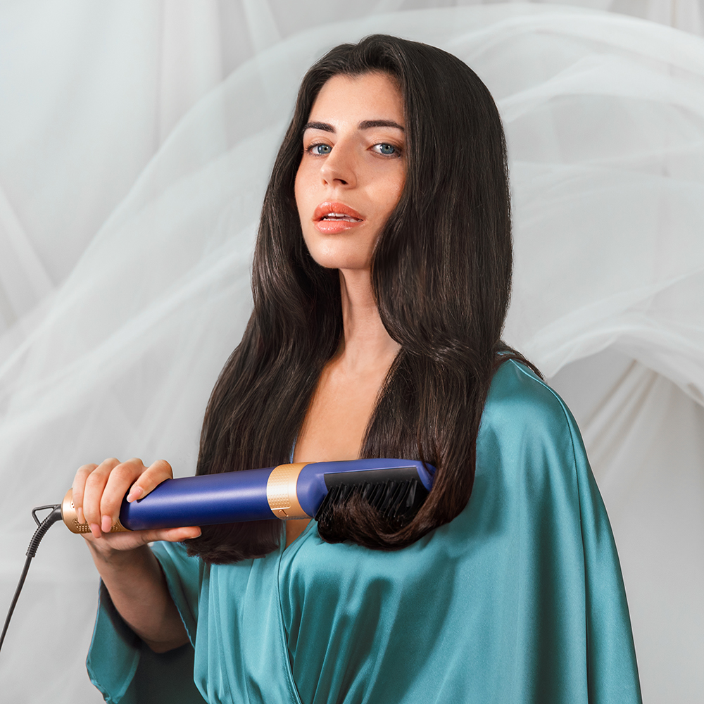 Mujer con cabello largo y oscuro peinándolo con un moldeador de cabello azul y dorado, usando una bata de satén azul.