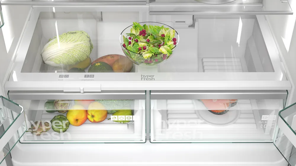 Offener Kühlschrank mit Gemüse, Salat und Obst. Oberes Regal mit Kohl und Salat. Unten Obst und Fisch.