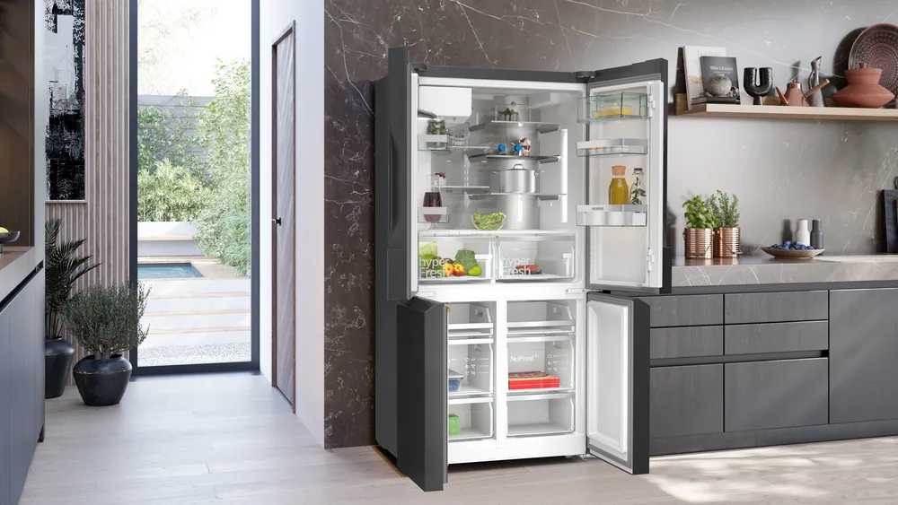 Offener Kühlschrank in einer modernen Küche. Obst, Töpfe und andere Gegenstände im Inneren. Tür zu einem Garten im Hintergrund.