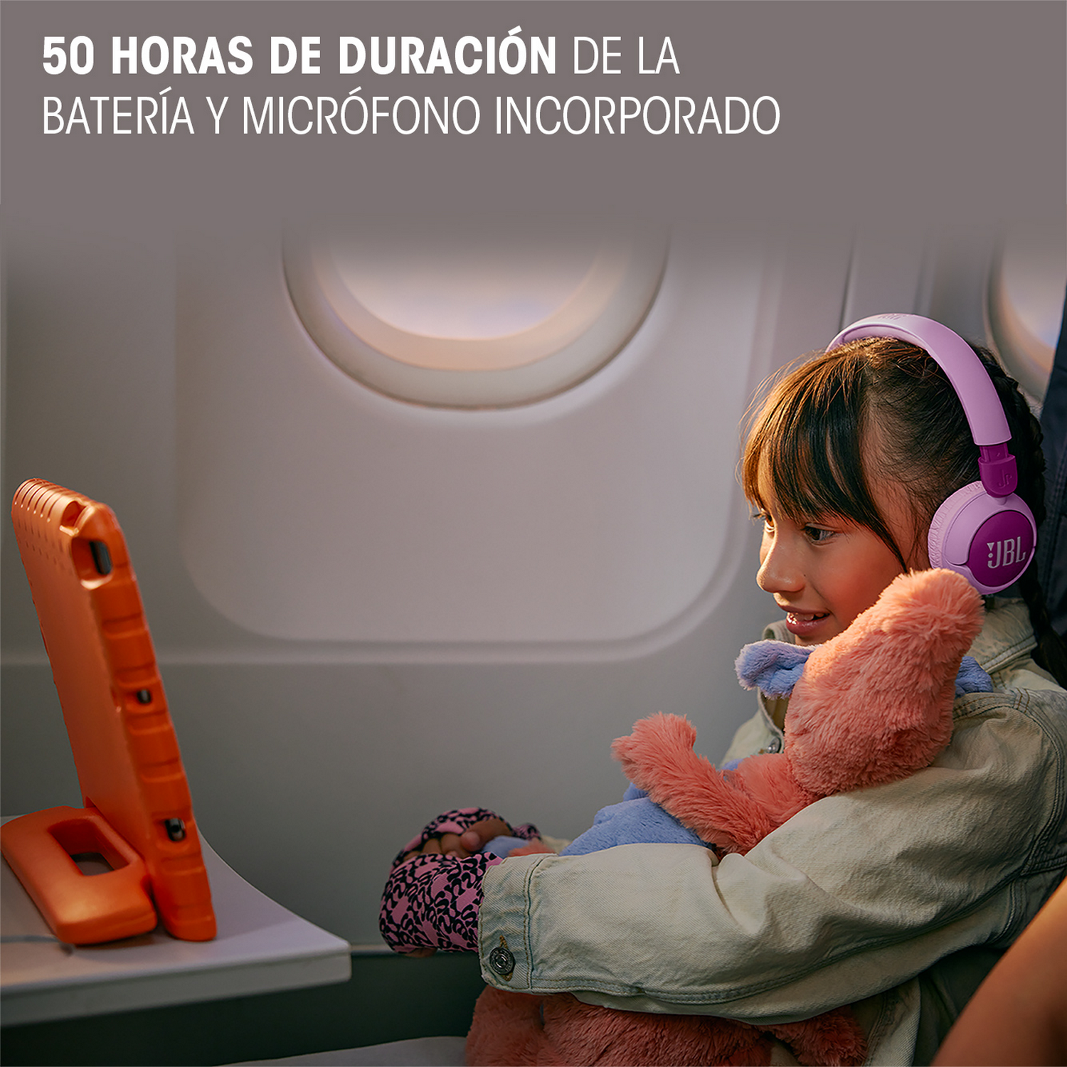 Una niña en un avión usa auriculares y mira una tableta. Una funda naranja está en la bandeja.