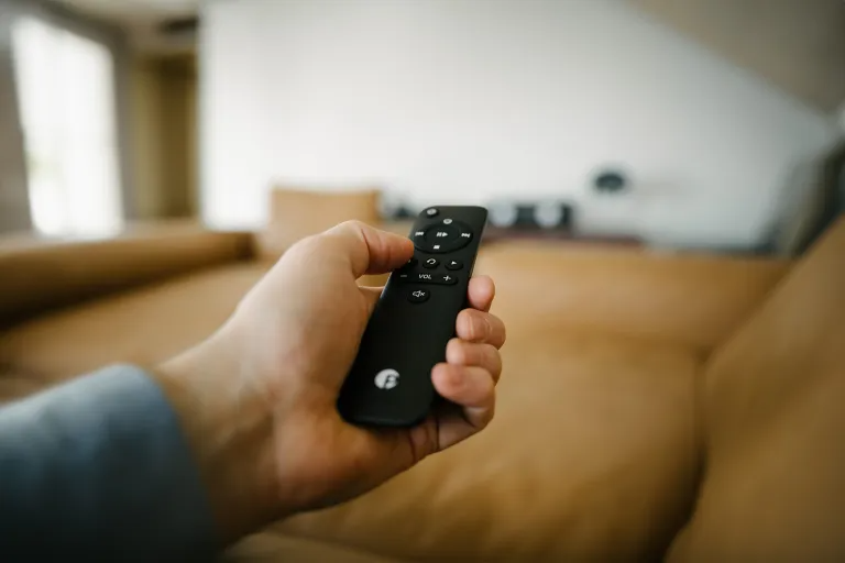 Hand hält eine schwarze Fernbedienung. Im Hintergrund eine braune Couch und eine weiße Wand.