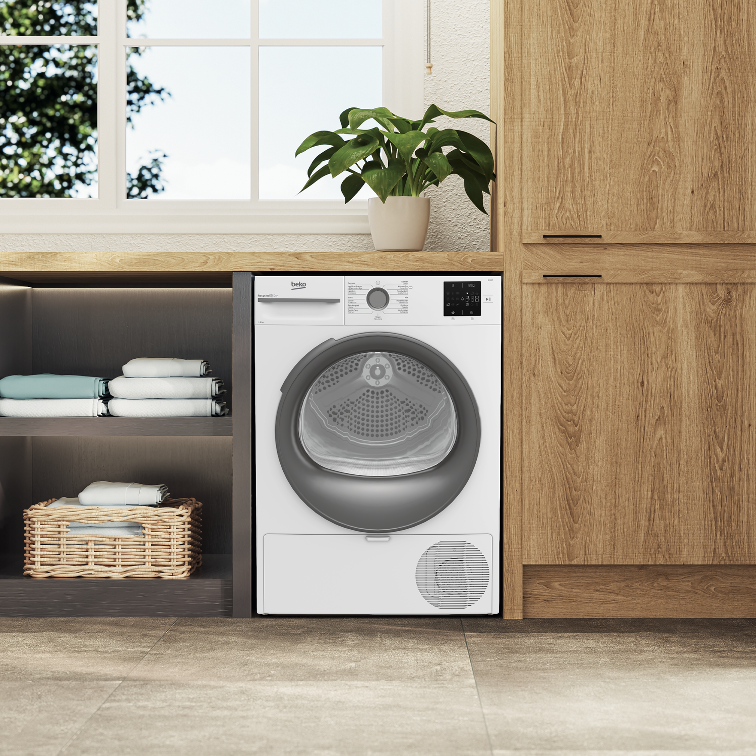 Een witte Beko droger in een wasruimte. Er is een raam, een plant en planken met opgevouwen handdoeken.