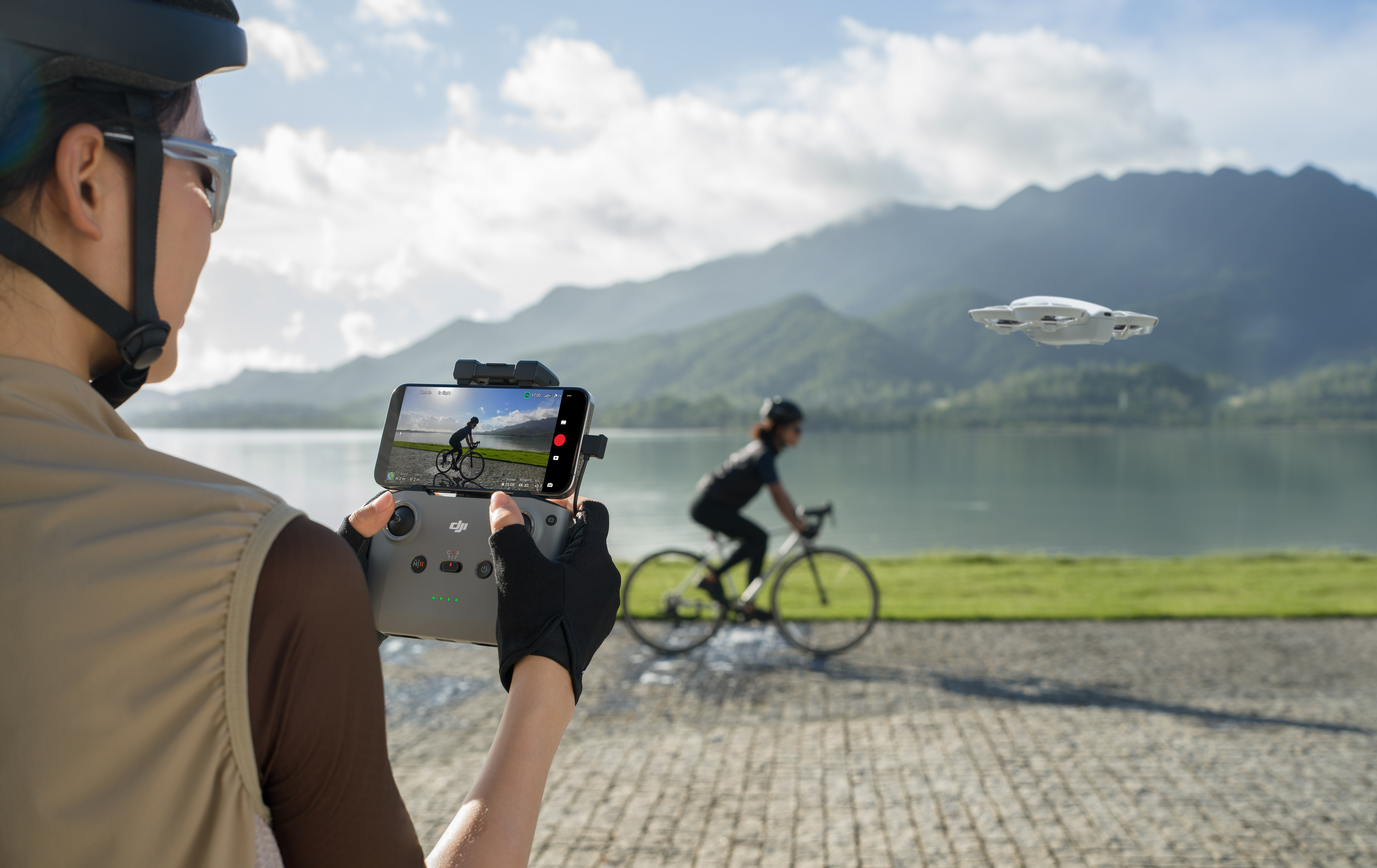 Mann mit Drohnen-Controller, filmt Radfahrer am See, Berge im Hintergrund.
