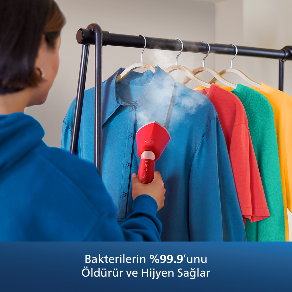Kişi, asılı kıyafetlere kırmızı bir buhar makinesi kullanıyor, metin %99,9 bakteri öldürür diyor.