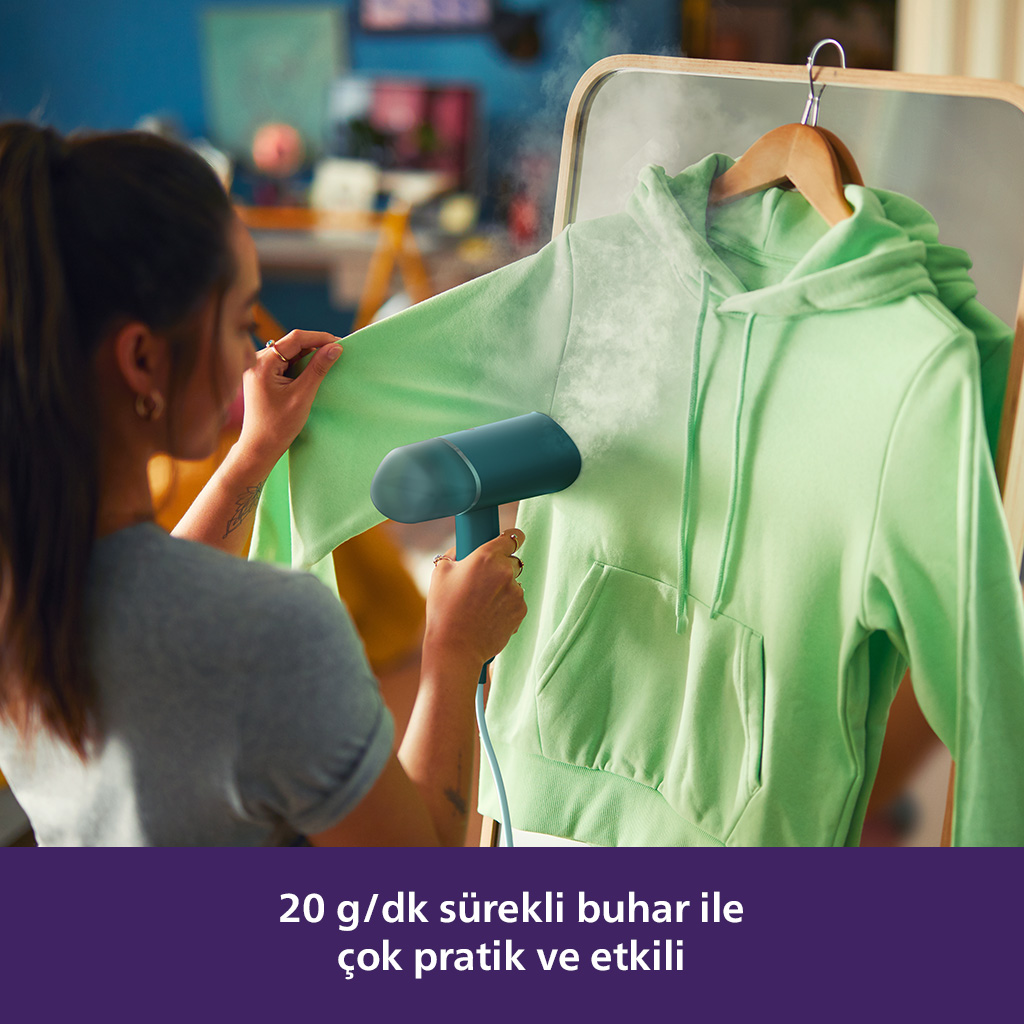 Kadın, yeşil bir kapüşonluyu el tipi bir buhar makinesiyle buharlıyor. Buhar görünür. Metin: 20 g/dk sürekli buhar.
