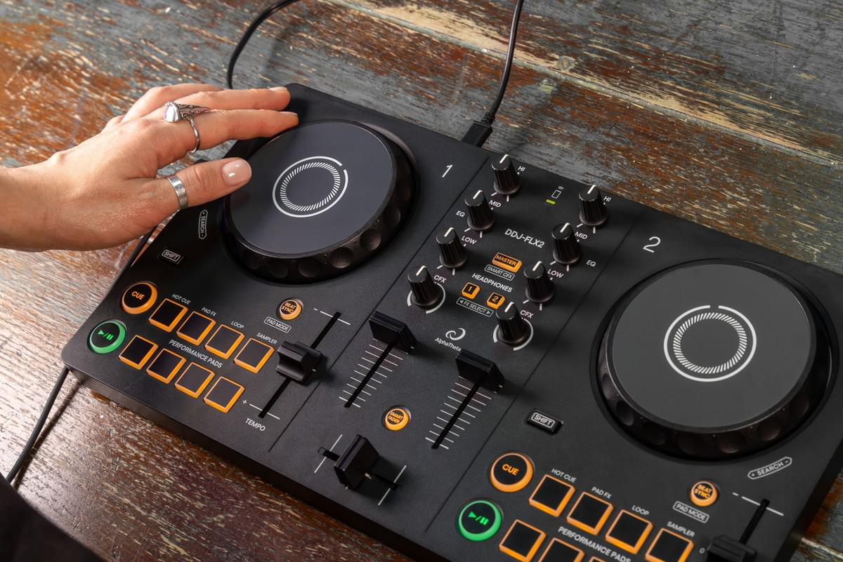 DJ-Controller, Hand am Jogwheel. Schwarze Oberfläche, Knöpfe und Drehregler sichtbar. Hintergrund: Holztisch.