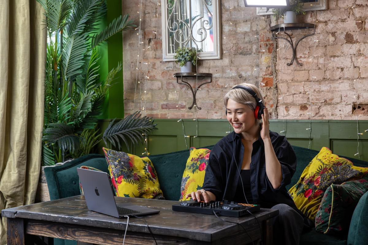 Frau mit Kopfhörern lächelt, benutzt einen Mixer mit einem Laptop auf einem Tisch.