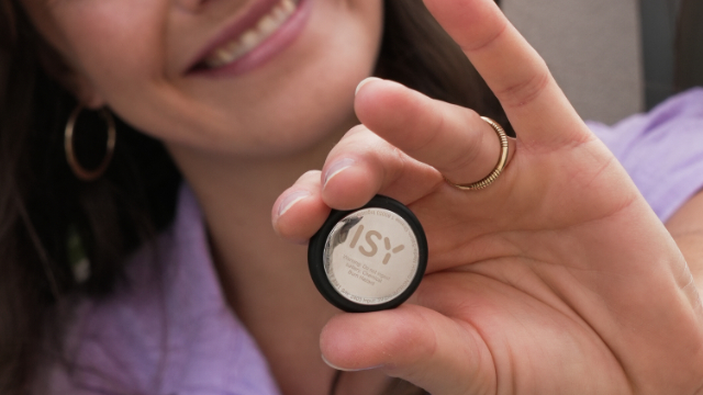Frau hält ein kleines, rundes, schwarzes Gerät. Das Gerät hat ein silbernes ISY-Logo und einen Ring. Hintergrund unscharf.