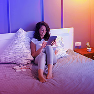 Eine Frau sitzt auf einem Bett und benutzt ein Tablet. Der Raum hat lila und orangefarbene Beleuchtung.