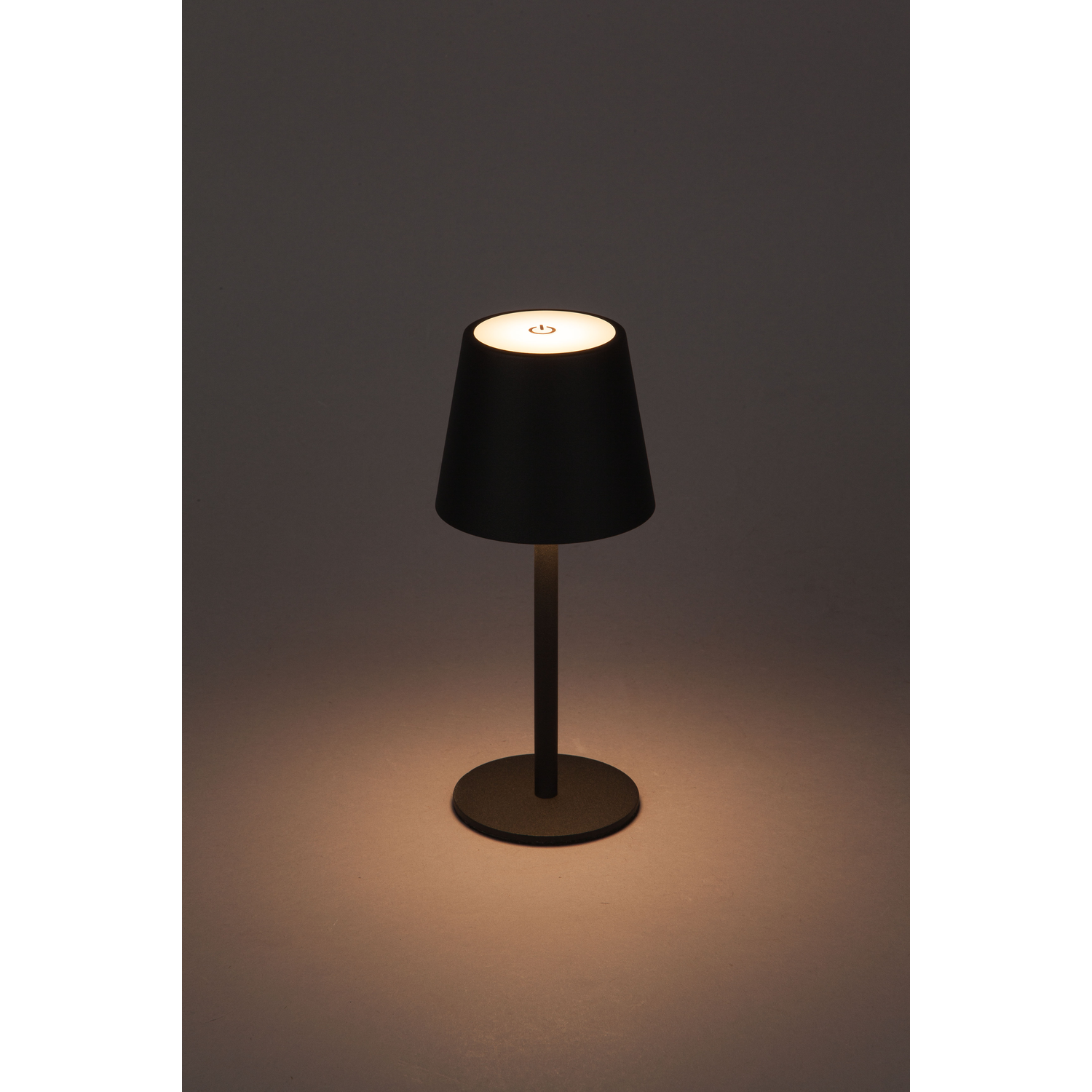 Zwarte tafellamp met een conische kap, die warm licht uitstraalt. De voet is cirkelvormig en donker.