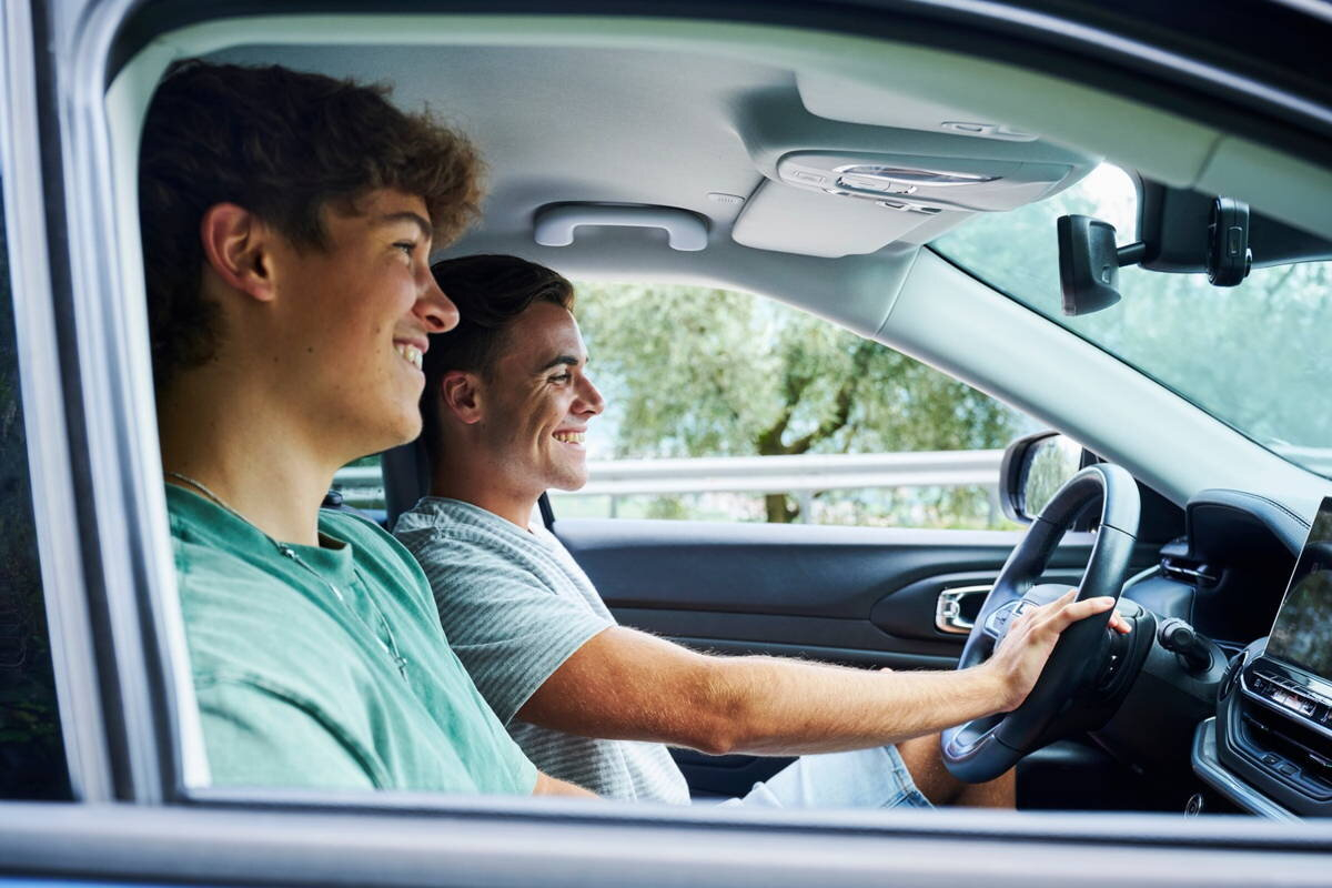 Dwóch młodych mężczyzn uśmiecha się w samochodzie. Jeden prowadzi, drugi siedzi na siedzeniu pasażera.
