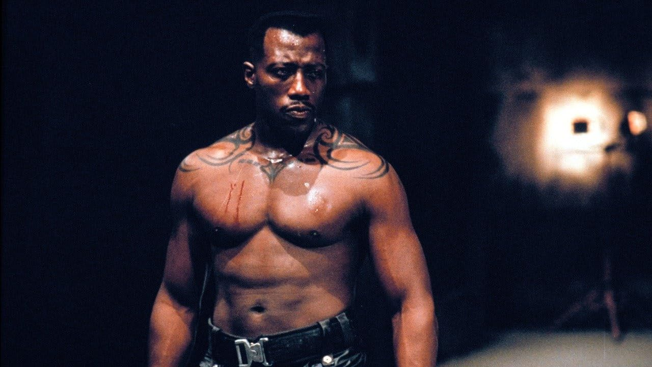 Un homme torse nu avec des tatouages. Il est musclé, debout dans un environnement sombre et granuleux.