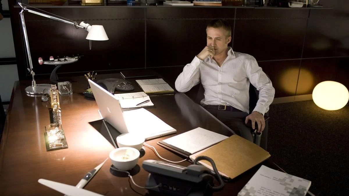 Homme assis à un bureau, l'air pensif. Ordinateur portable, lampe et documents sur le bureau.