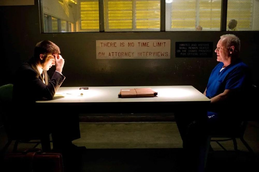 Deux hommes à une table dans une pièce sombre. Un homme est en costume, l'autre en blouse bleue.