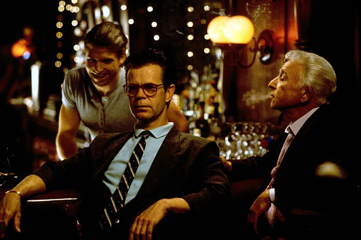 Drie mannen in een bar. Een man met bril en stropdas zit op de voorgrond. Een ander rookt. Een derde glimlacht.