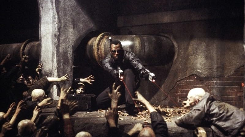 Blade, dans un cadre sombre, combattant avec deux armes, entouré de zombies.