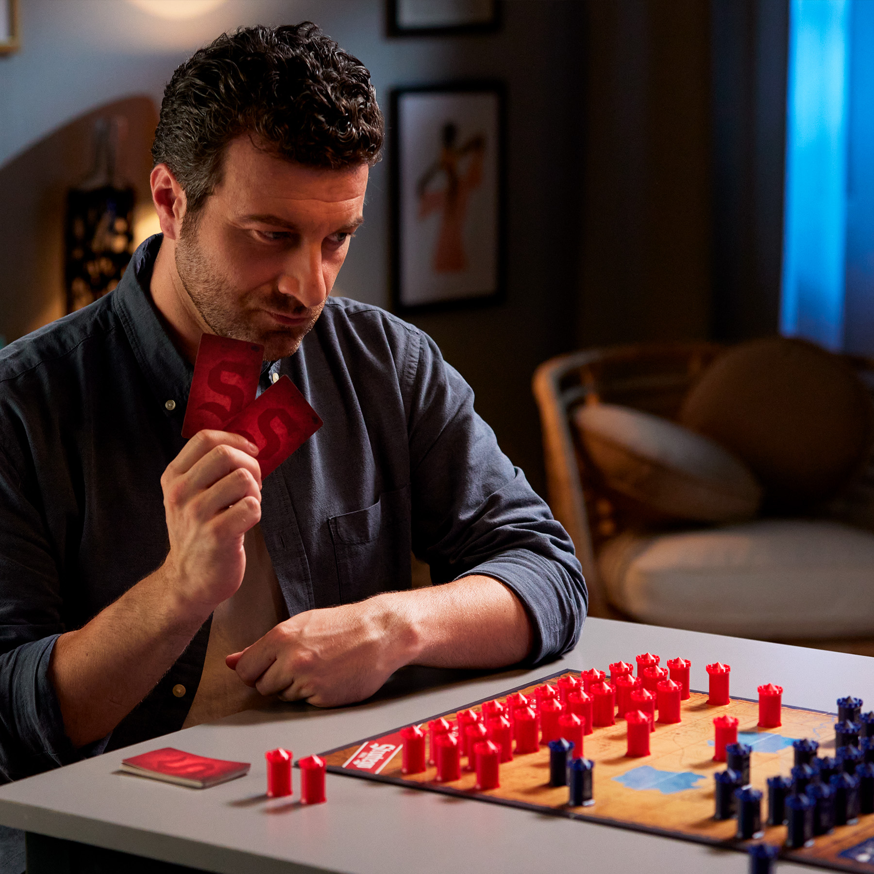 Een man die kaarten vasthoudt, kijkt naar een bordspel met rode en blauwe stukken.