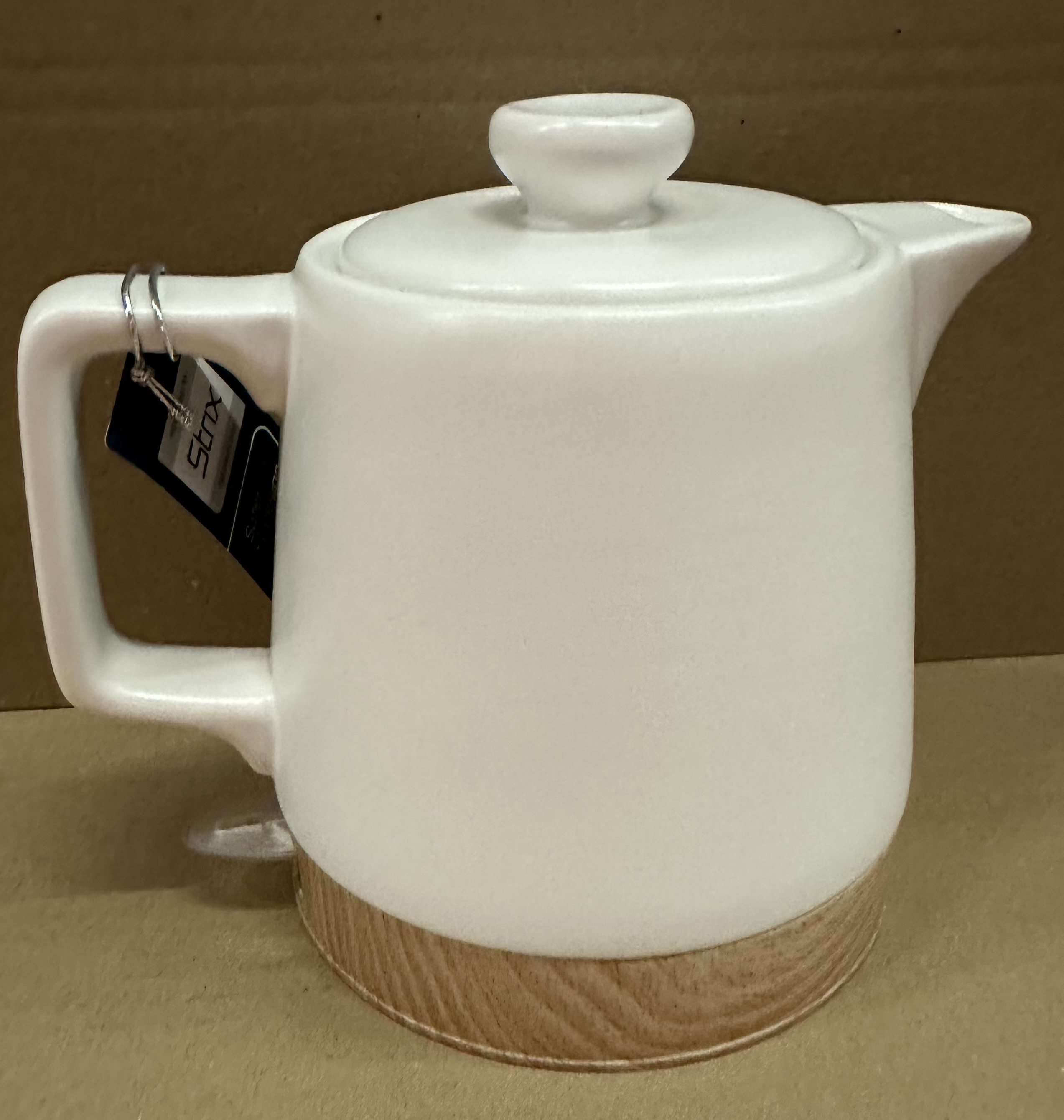 Biały ceramiczny czajnik z pokrywką i drewnianą podstawą. Uchwyt jest z boku.