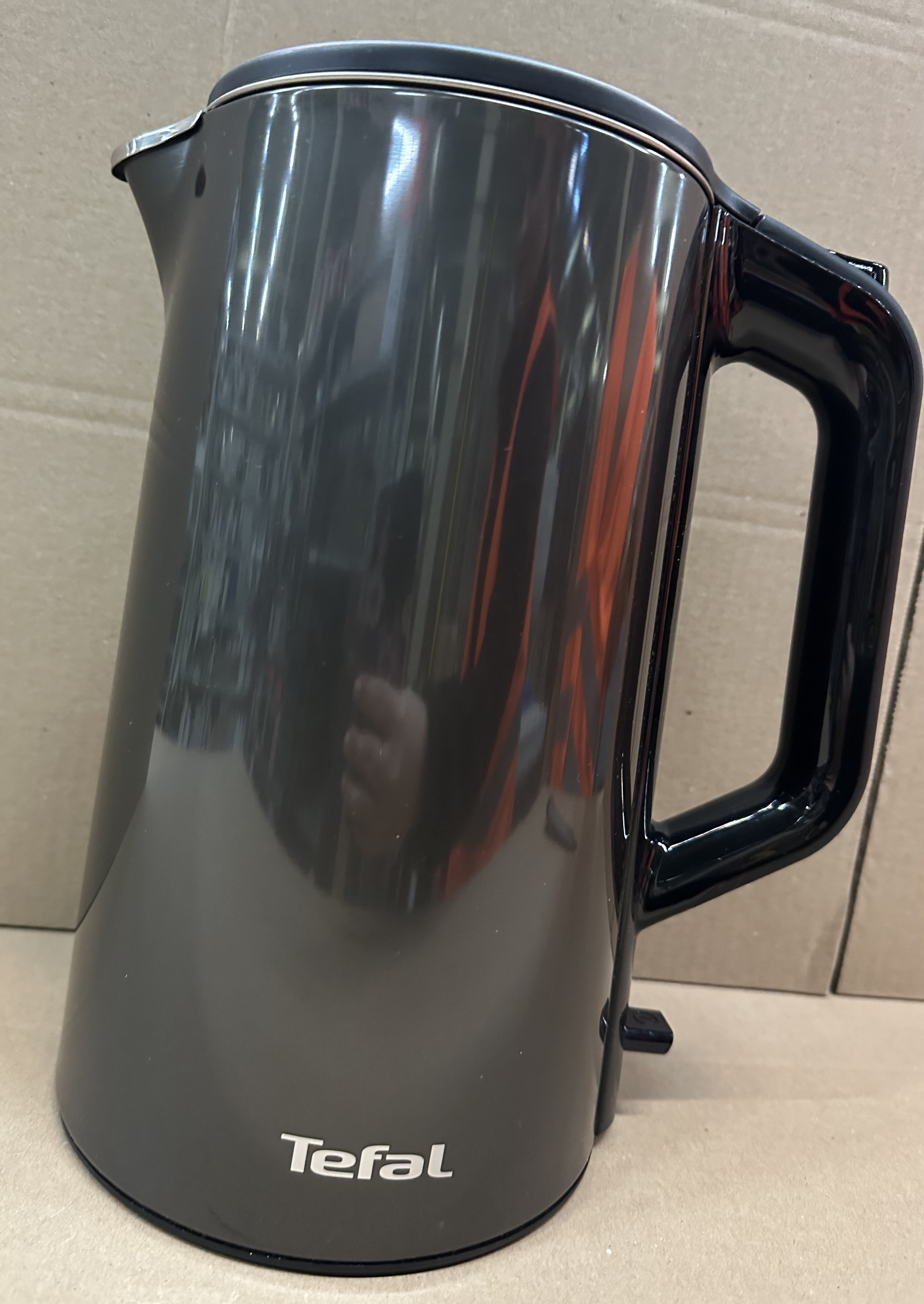 Ciemny, błyszczący czajnik z czarnym uchwytem stoi na kartonie.