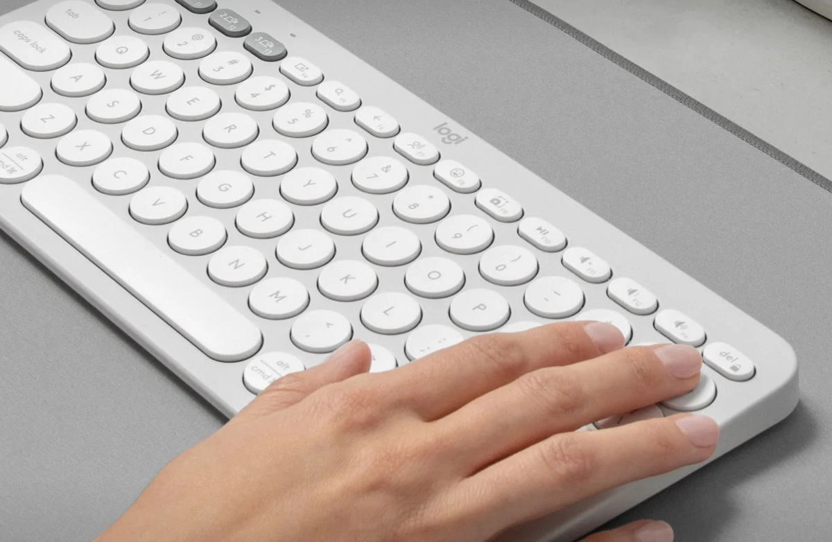 Közelkép egy kézről, mely egy fehér Logitech billentyűzettel érintkezik szürke felületen.