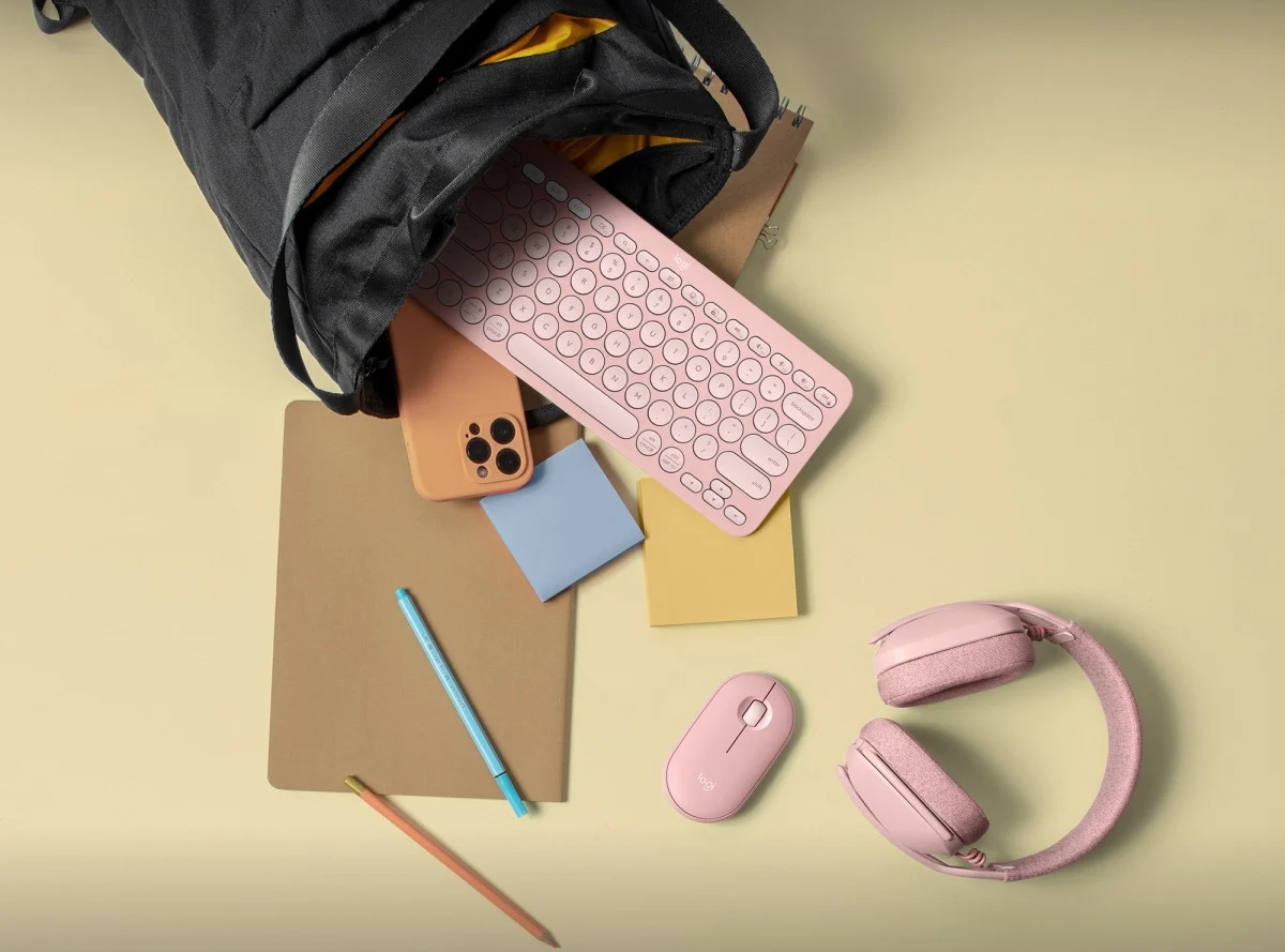 Egy fekete táska rózsaszín billentyűzettel, telefonnal, egérrel és fejhallgatóval. Van egy jegyzettömb és egy ceruza is.