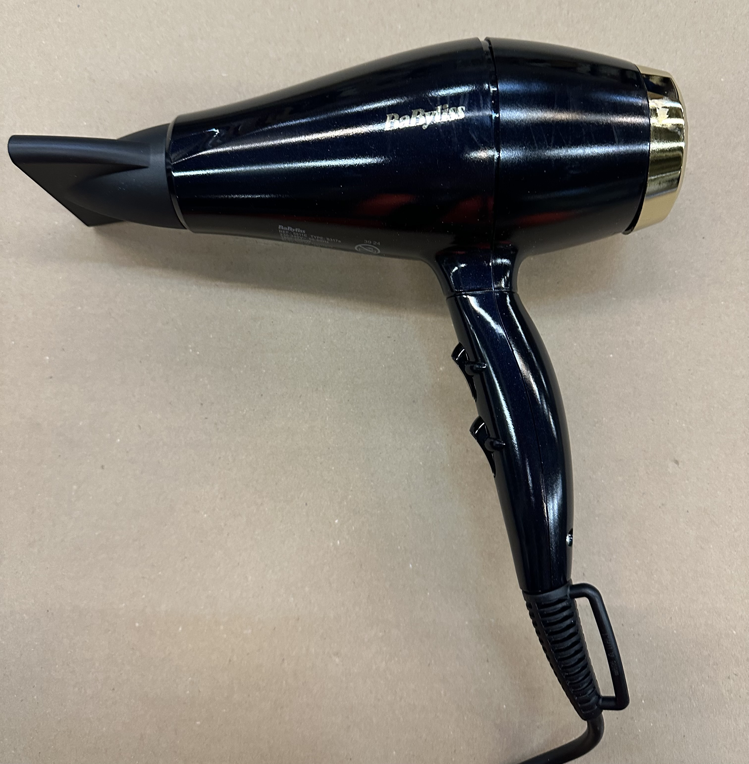 Błyszcząca, czarna suszarka do włosów ze złotym akcentem na rączce spoczywa na jasnobrązowej powierzchni.